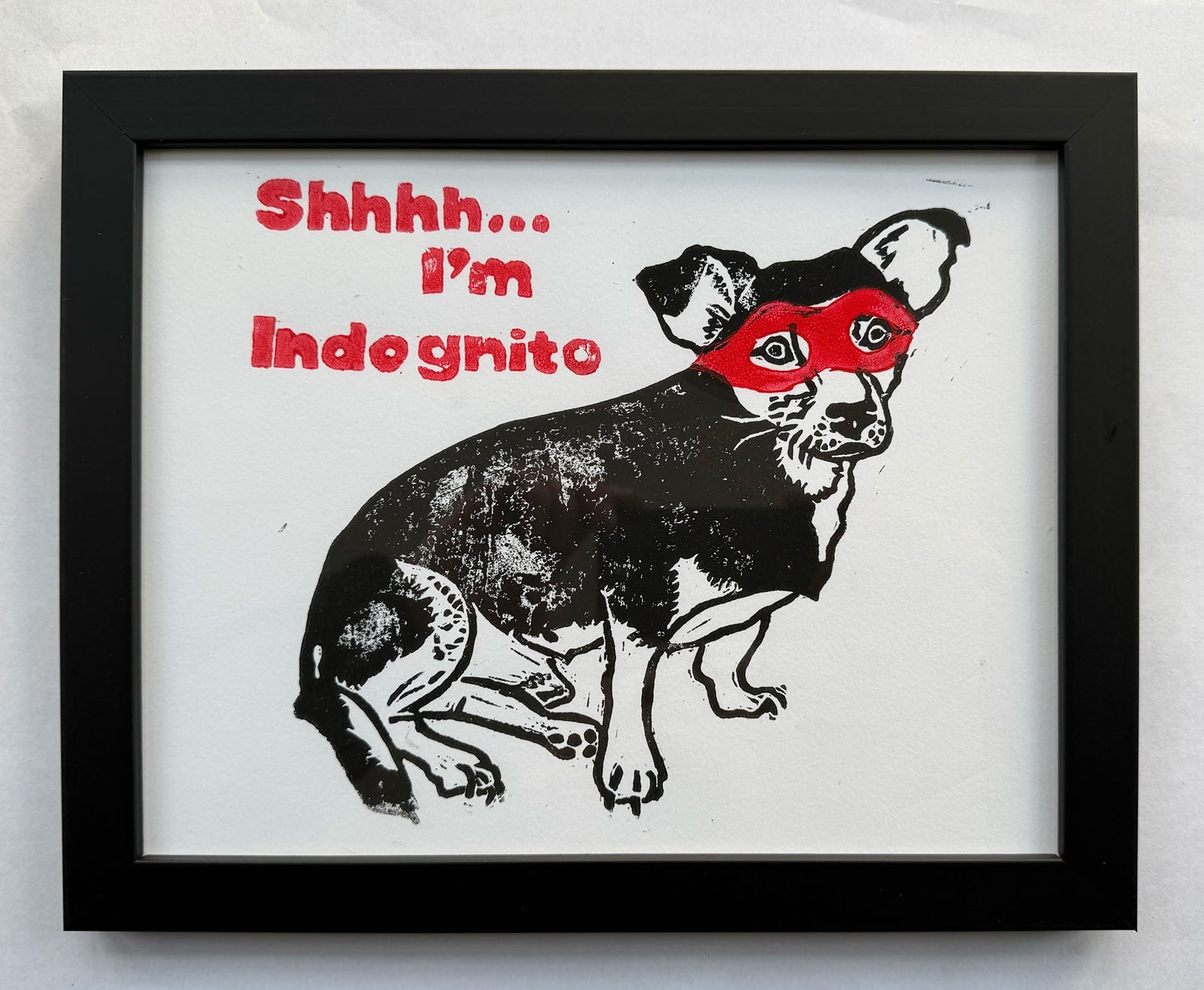In Frame image of a hand-printed two-colour linocut featuring a small dog in a red eye mask.